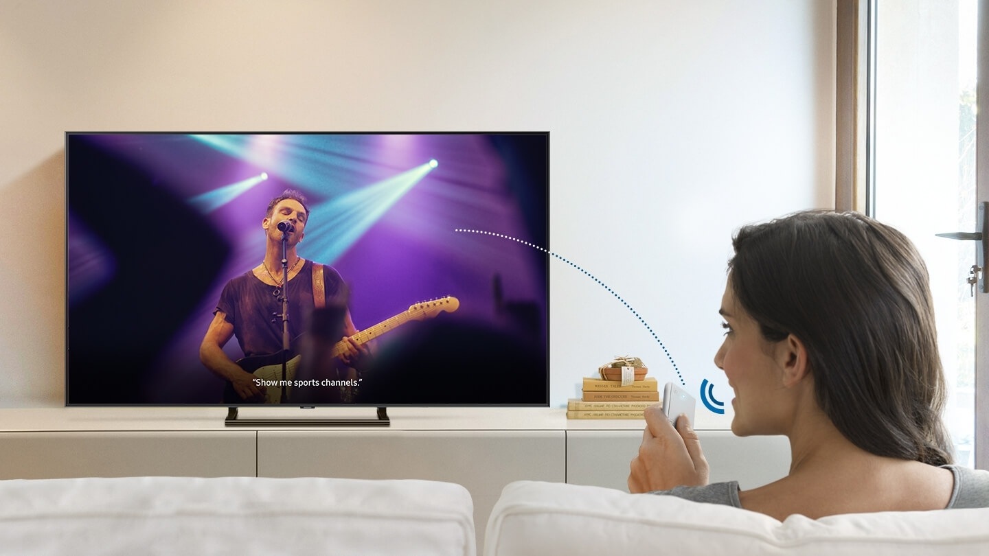 A woman is using a voice assistant to ask the title of the song playing on Smart TV, through the One Remote Control.