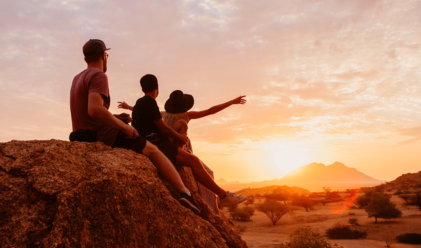Une famille assise sur des rochers contemple la beauté de la nature en regardant un coucher de soleil au loin sur une montagne.