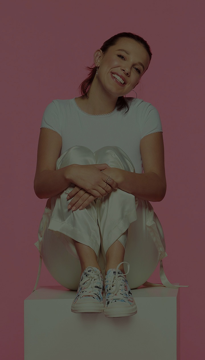 Portrait studio de Millie Bobby Brown sur un fond rose. Souriante, la tête penchée sur le côté, elle est assise sur une boîte blanche et croise ses mains pour tenir ses jambes.