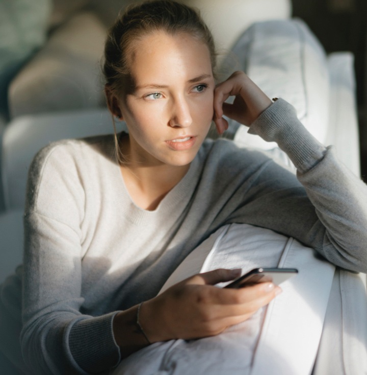 Uma mulher com uma camisola bege, sentada no sofá, segura o seu telefone Galaxy. Tem o corpo está voltado para a parede e olha pela janela com a cabe?a apoiada na m?o esquerda.