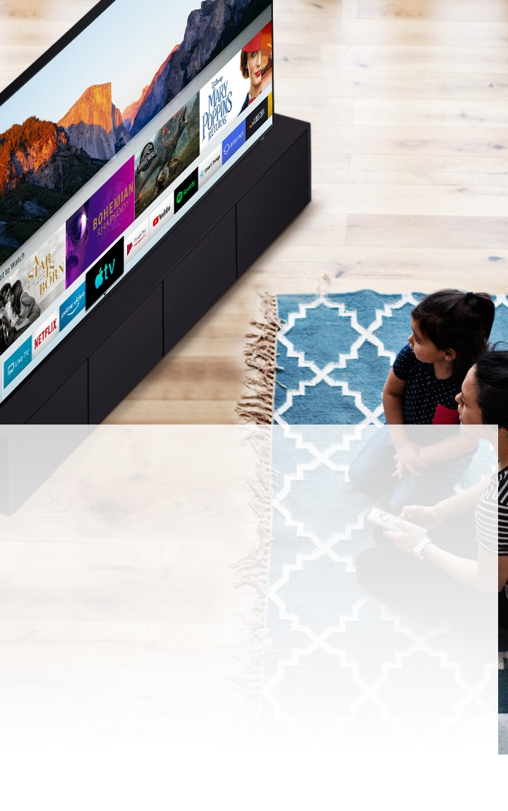 Una madre y una hija sentadas en una alfombra azul con patrones blancos frente a un televisor inteligente Samsung. Están mirando las diversas aplicaciones instaladas en un televisor inteligente.
