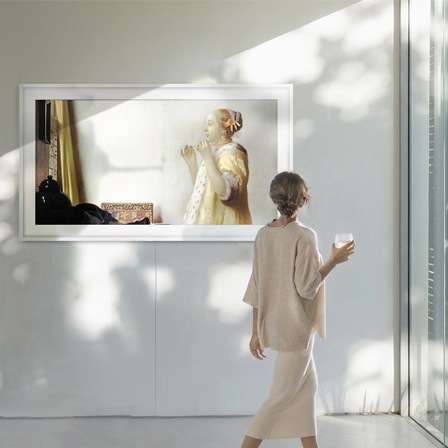The Frame hängt an einer Wand und zeigt ein Gemälde von Johannes Vermeer, 'Junge Dame mit Perlenhalsband', das aus dem Wohnzimmer eine Kunstgalerie macht. Eine Frau schaut sich das Kunstwerk an.