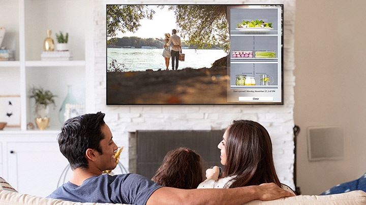 The family sitting on the couch and watching TV hanging on the wall. They are watching TV video screen and a part of the inside of a refrigerator are coming out on TV.