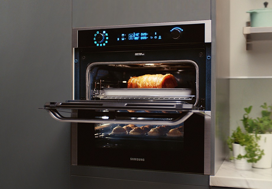 The Dual Cook Flex™ (NV7000N) oven is shown at a tilt, installed into a grey wall. The top half of the Flexible Door is open. At the top of the oven is a digital dashboard which shows from left to right, the Smart Control, oven light, temperature indicating 210 degrees in the upper cavity and 180 degrees in the lower cavity, oven timer, and control knob. A roast beef is cooking in the upper cavity while muffins are cooking in the lower cavity.