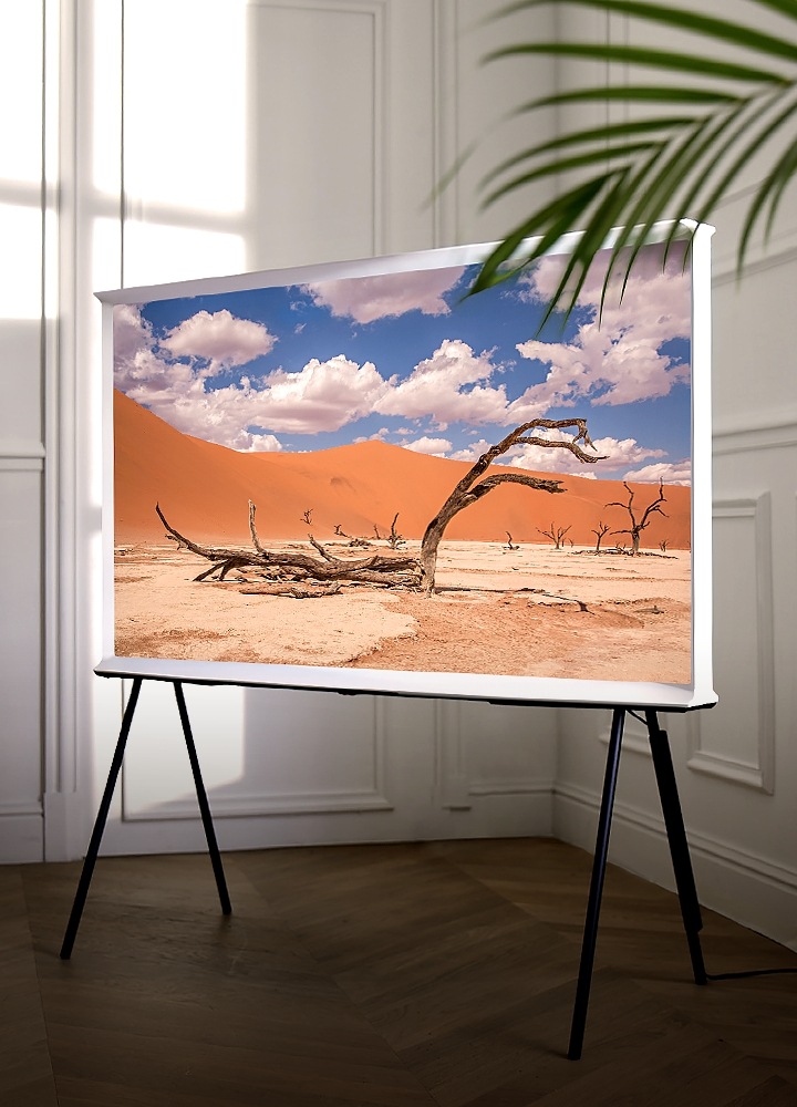 QLED-technologie KV Een Cloud White-versie van The Serif staat op een TV-standaard in de hoek van een kamer, met een schitterend beeld van een woestijn met een blauwe lucht en witte wolken.
