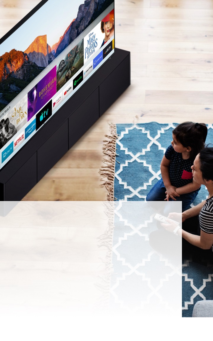 Een moeder en dochter zitten op een blauw vloerkleed met een wit patroon voor een Samsung Smart TV. Ze kijken naar de verschillende apps die op de Smart TV zijn geïnstalleerd.