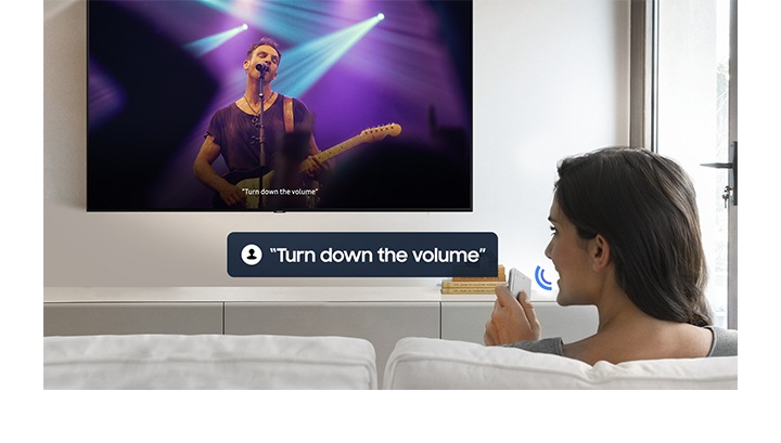 Een vrouw zit op een witte bank in een helder verlichte kamer en kijkt naar een video van een concert op een Smart TV die aan de muur hangt. De vrouw activeert de spraakassistent en geeft de afstandsbediening van de Smart TV het commando ''Turn down the volume'' om het geluid zachter te zetten.