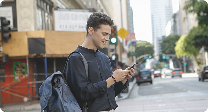 Un homme avec un sac à dos est débout au bord de la rue et regarde son téléphone qu’il tient des deux mains.