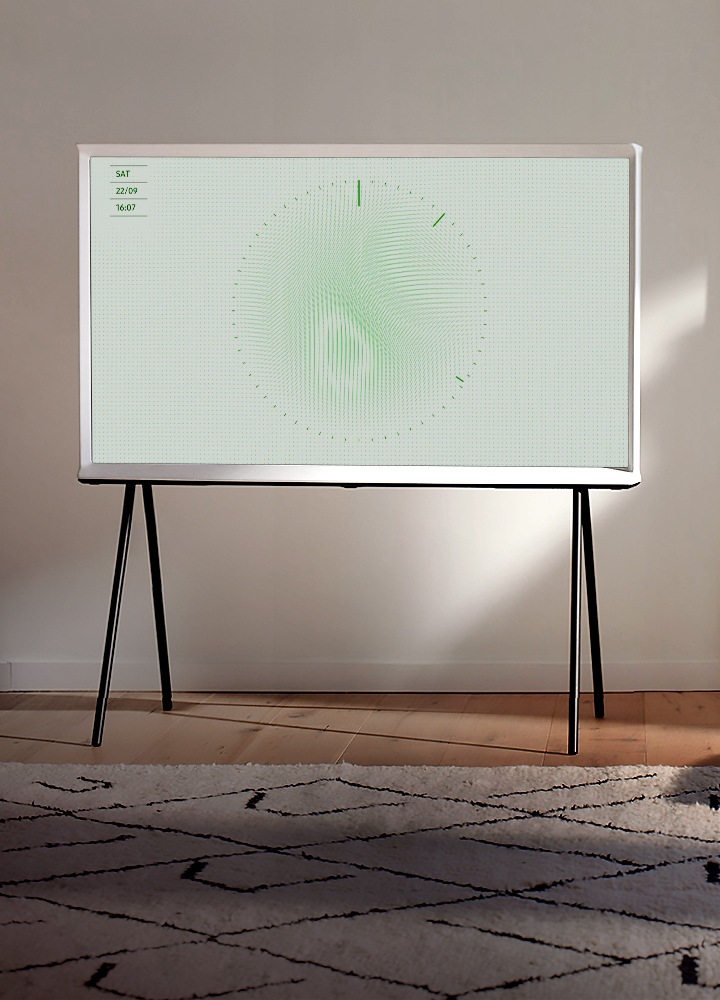 Design KV Un modèle White de la Serif est posé sur un support noir dans une pièce de séjour claire et élégante, avec un tapis décoratif, un fauteuil vert et élégant à trois pieds et de larges fenêtres. L’image à l’écran est le Leaf Ambient Mode.