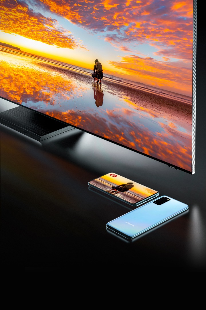Samsung QLED e dois Galaxy S20 colocados abaixo da tela da TV. Os produtos compartilham o mesmo conteúdo de 8k de uma pessoa agachada em um deserto salgado refletindo o céu vermelho.