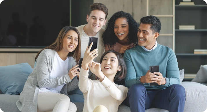 Um grupo de amigos está sentado no sofá enquanto a mulher do meio está agachada no chão e segurando seu smartphone para tirar uma selfie do grupo.