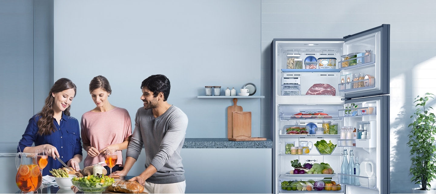 Geladeira Samsung Twin Cooling Plus aberta com frutas, legumes e garrafas de água em uma cozinha com um homem e duas mulheres sorrindo