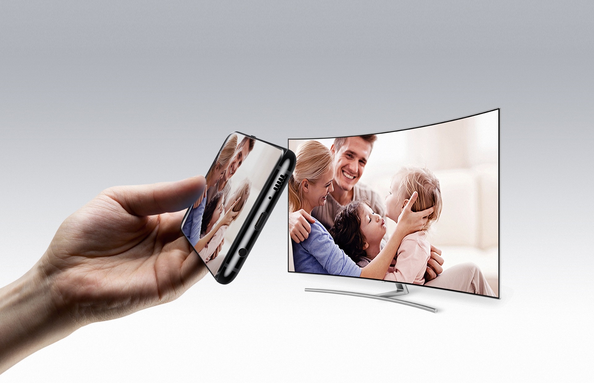 Uma mão segura um smartphone Samsung ao lado esquerdo da imagem. Na tela do celular vemos uma foto de pai, uma mãe e dois filhos. Ao lado direito da imagem, a mesma foto aparece em uma TV Samsung 4k.