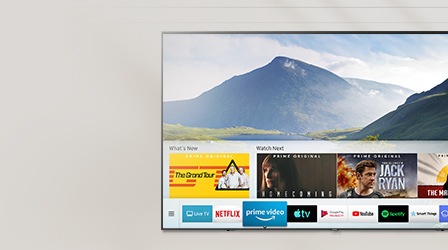 A interface de usuário do Smart Hub é exibida sobre um plano de fundo de natureza na tela da TV; a interface de usuário contém vários conteúdos, como “O que há de novo”, “Assista a seguir” e vários aplicativos como Netflix, Prime Video, Apple TV, Google Play Filmes e TV, YouTube, Spotify, SmartThings, Internet, DEEZER.