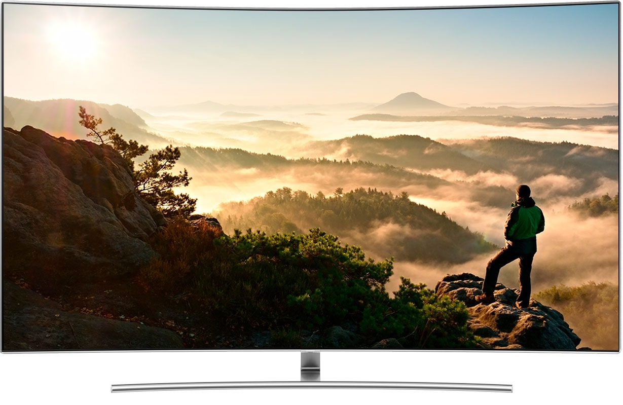 Animação na tela da Smart TV Samsung em que vemos um cara olhando para uma paisagem de montanhas rochosas e sobem os ícones do Smart Hub e o Netflix fica em destaque. Após isso, vemos outra imagem na tela, de uma cena da série The Crown, com a atriz que interpreta a Rainha do Reino Unido vestida de azul, acenando para um grupo de pessoas.