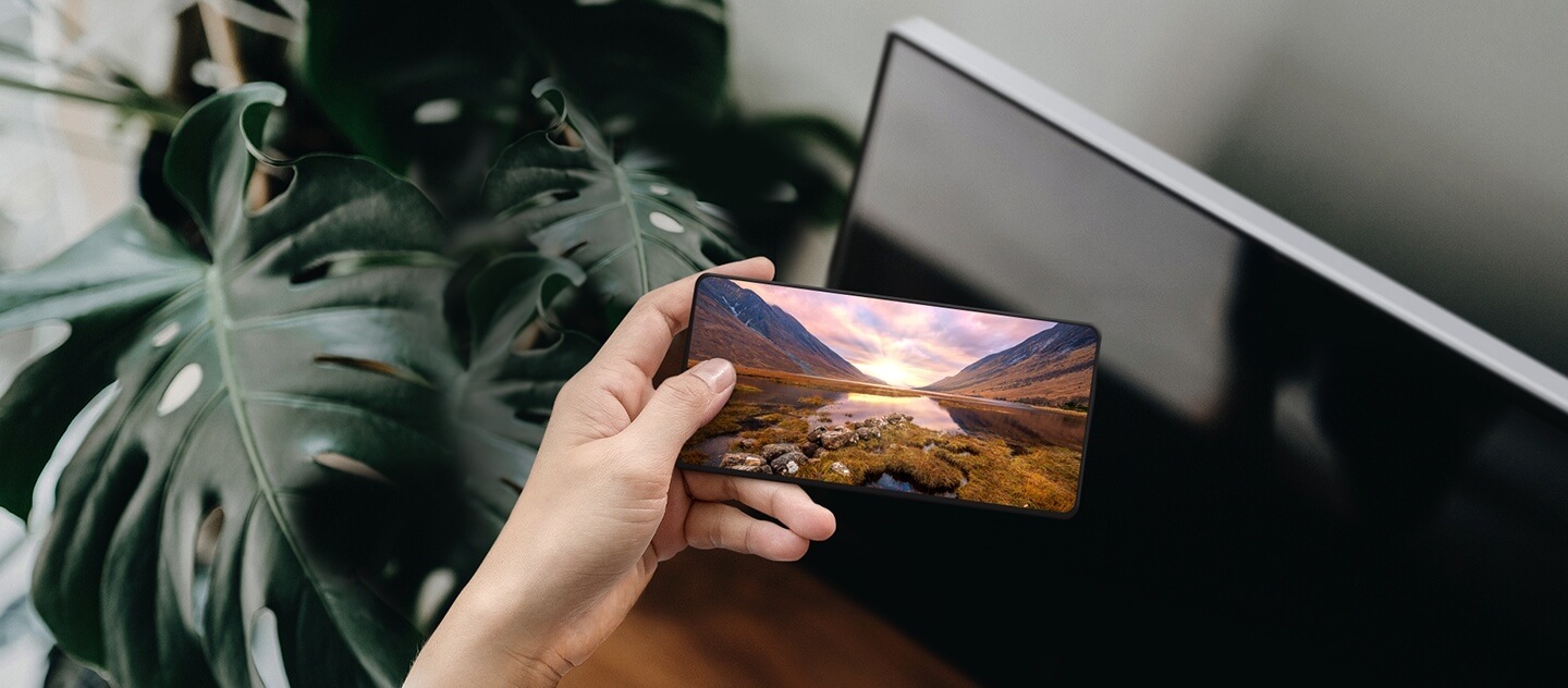 A hand holding a Galaxy smartphone in front of a Samsung TV. The Galaxy screen shows a majestic landscape image.