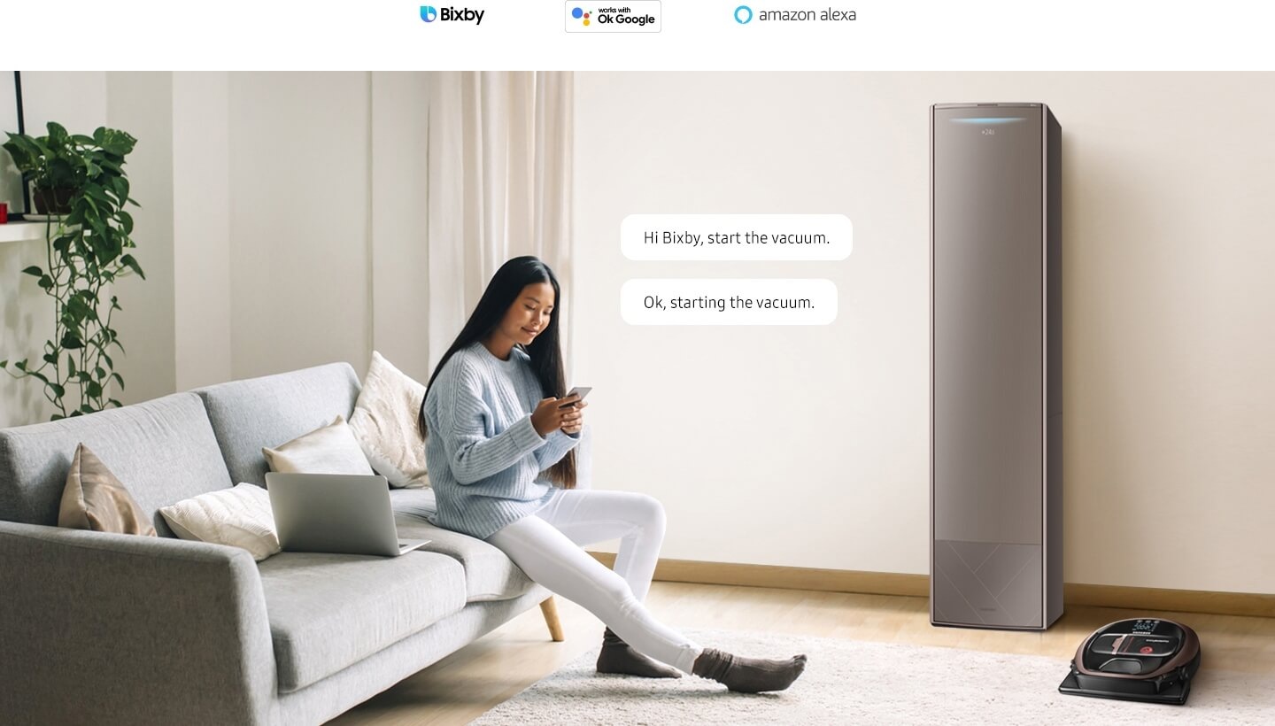 A woman sits on her sofa with her smartphone. There is a smart air conditioner and a smart robot vacuum in the room. 2 conversation bubbles appear near the woman with the text "Hi Bixby, start the vacuum" and "Ok, starting the vacuum". 3 AI assistant logos are displayed above: Bixby, Hey Google, and Amazon Alexa.