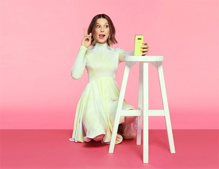 Millie Bobby Brown kneels while holding up a peace sign with her fingers as she takes a selfie with a neon yellow Samsung Galaxy A80 that's propped on a white stool.