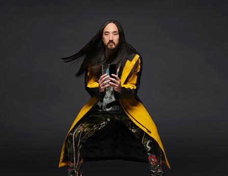 Steve Aoki standing with his knees slightly bent outward while sporting a yellow long coat as he holds up an unfolded Samsung Galaxy Fold smartphone
