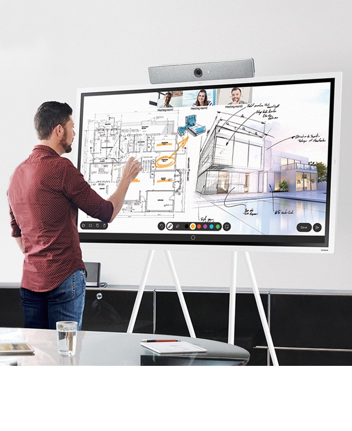 A man is working on the Samsung Flip monitor, with a video camera at the top of the monitor. At the top of the monitor there are three people in a video conference. The screen shows blueprints for architectural plans and a rendering of the house.