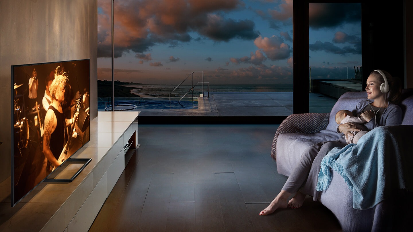 A woman is watching TV in the living room with a headset while holding a baby at night.
