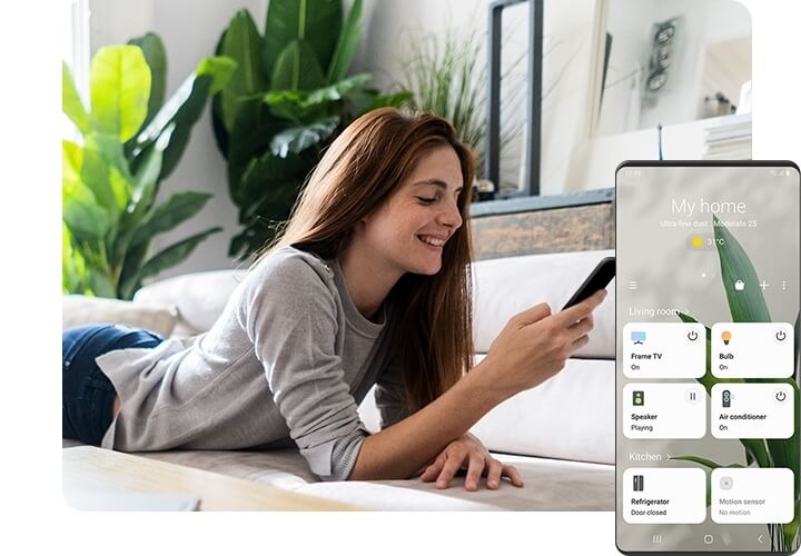 Une femme souriante est étendue sur le divan et regarde son téléphone intelligent. Du côté droit, à l’avant-plan, un écran Galaxy affiche l‘IUG de l’appli SmartThings avec des options de commande pour divers appareils domestiques dans le salon, le niveau de qualité de l’air et la température courante.