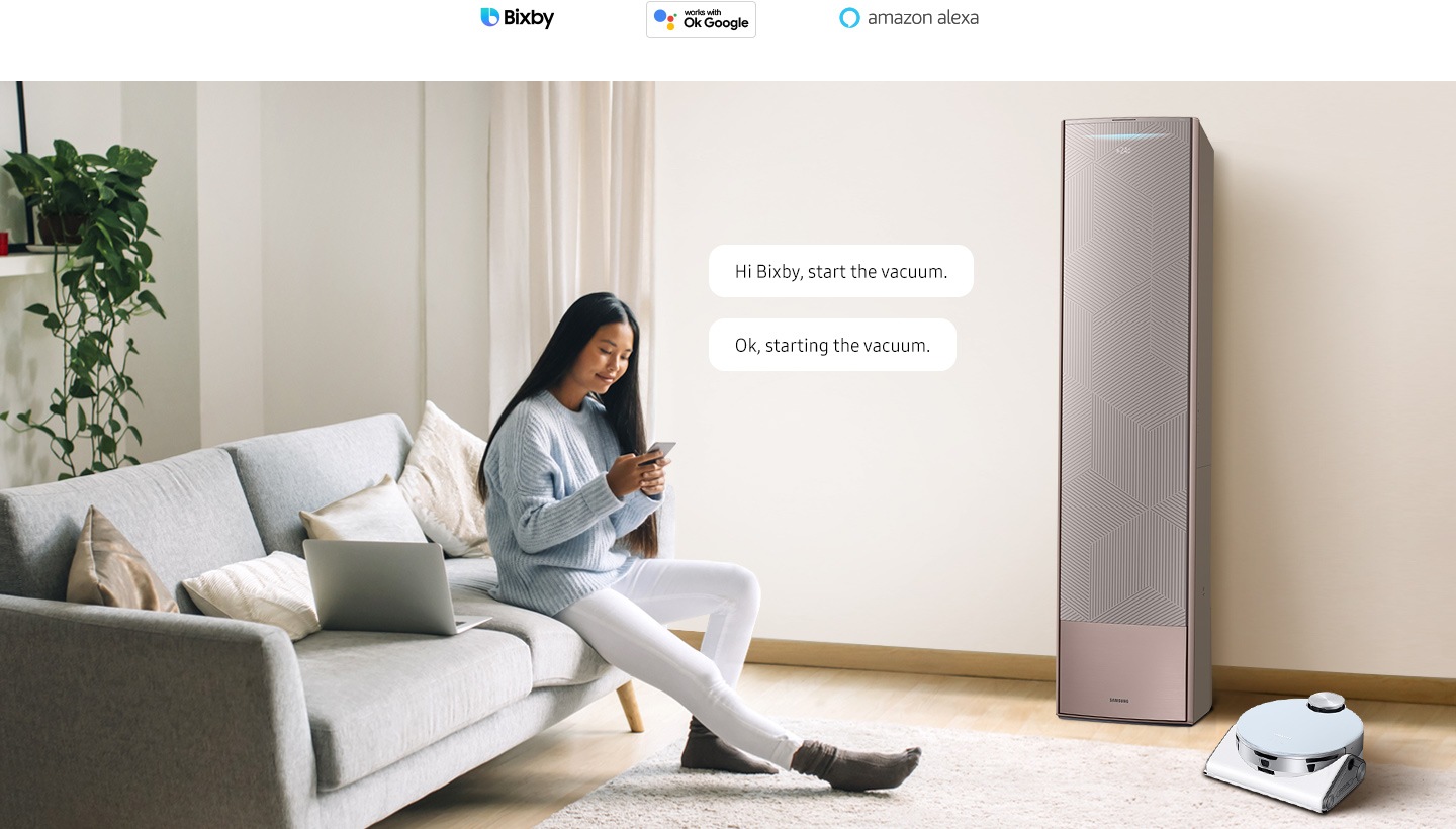 Une femme prend place sur son divan avec son téléphone intelligent Il y a un climatiseur et un robot aspirateur intelligents dans la pièce. Deux bulles de texte s’affichent près de la femme, une contenant le texte « Bonjour Bixby, démarre l’aspirateur » et l’autre « Ok, démarrage de l’aspirateur ». Trois logos d’assistance IA sont affichés ci-dessus : Bixby, Hey Google et Amazon Alexa.