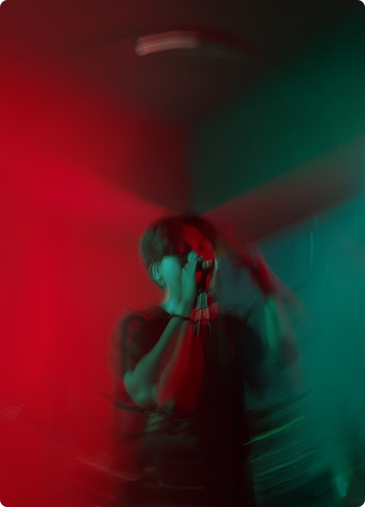 why-galaxy-powerful-performance Un chanteur est debout sur une scène fortement éclairée, avec un éclairage rouge à gauche et un éclairage vert à droite. L’image est légèrement brouillée, montrant du mouvement.