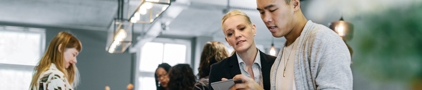 Une vue intérieure d’une boutique expérience+ Samsung remplie de gens. Au premier plan, un jeune homme montre son téléphone intelligent à une employée de la boutique qui est derrière son épaule.