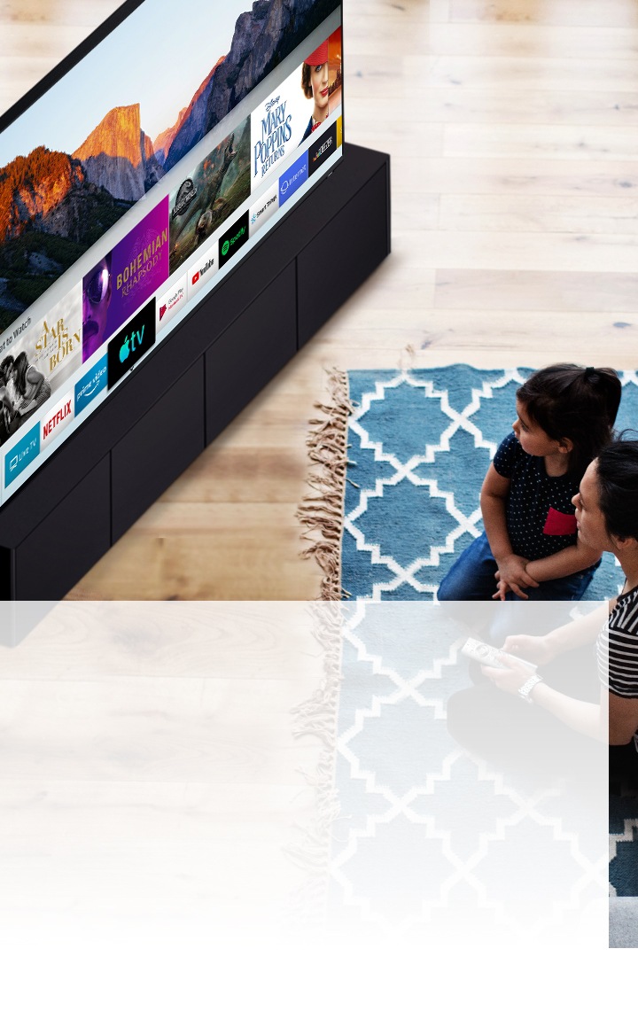 Une mère et sa fille assises sur un tapis bleu avec des motifs blancs devant un téléviseur intelligent Samsung. Elles regardent les différentes applications installées sur un téléviseur intelligent.