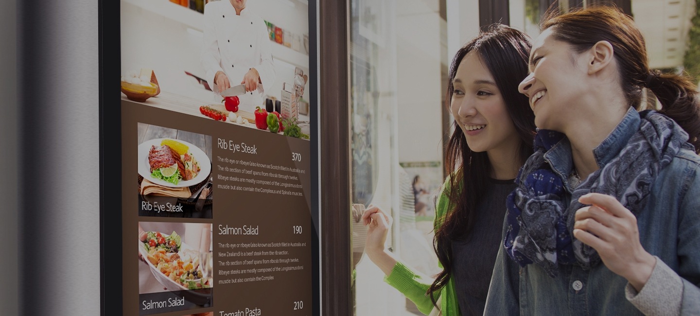 Zwei Frauen, die sich auf einem Samsung Outdoor Signage-Display-Bildschirm, der vor einem Restaurant aufgestellt ist, über das Menüangebot informieren