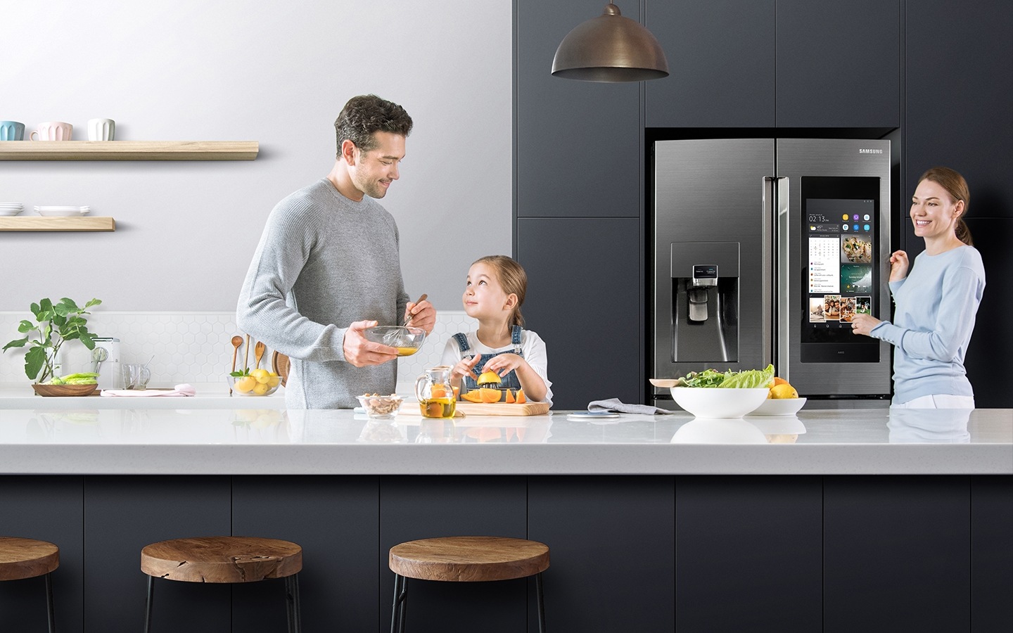 Ein Vater und eine Tochter bereiten Eier und Orangensaft auf der Küchentheke zu, während die Mutter über sie wacht und lächelt, während sie neben dem Family Hub mit dem Bildschirm im On-Modus steht.