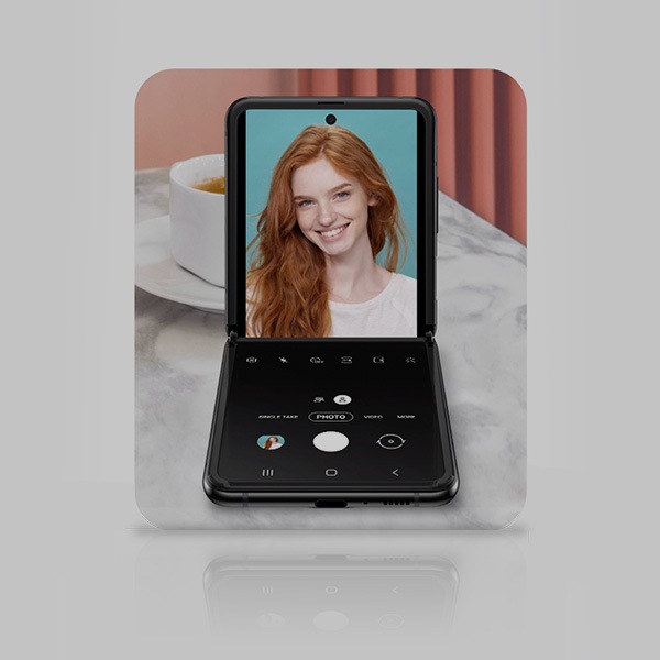 Ein schwarzer Galaxy Z Flip wird im Flex-Modus offen auf einem Marmortisch abgestützt. Die native Kamera-App wird im Fotomodus mit einer rothaarigen, lächelnden Frau im Display geöffnet.
