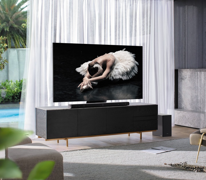 Das Bild zeigt einen Samsung QLED TV mit einer Samsung Soundbar, welche auf einem schwarzen Sideboard in einem modernen Wohnzimmer stehen. Auf dem Fernseher ist ein Bild einer Ballettänzerin die eien Übung auf einem schwarzen Boden macht.