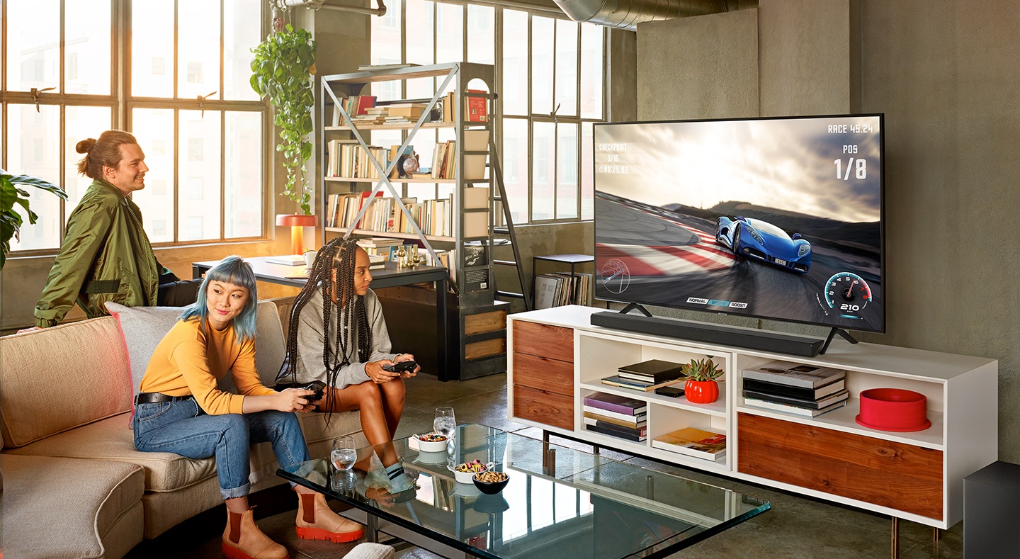 Una sala con tres personas disfrutando el momento mientras juegan y videojuego de carreras en un televisor Samsung QLED con barra de sonido