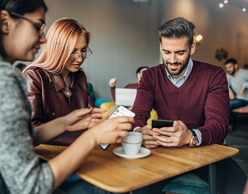 Im verdunkelten Hintergrund sieht man eine Gruppe von Männern und Frauen, die mit ihren Smartphones beschäftigt sind. Im Vordergrund zeigen zwei Galaxy Smartphone-Bildschirme, wie Private Share die Bildschirmerfassung von sensiblen Informationen wie Kreditkartendetails verhindert.