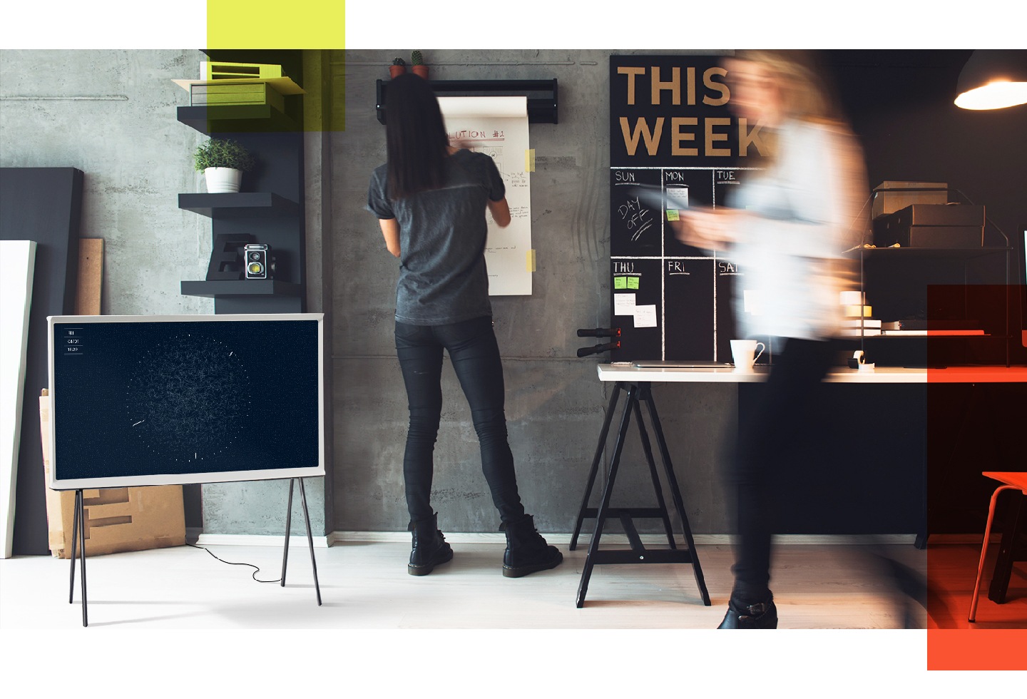Perfekte Harmonie foto 6 Ein weißer Serif TV im Ambient Mode mit Textil-Hintergrund mit einer marineblauen Grafik auf dem Display wird auf einem Standfuß in einem Büro gezeigt. Daneben ist eine Frau zu sehen, die etwas auf eine Tafel schreibt, während eine andere Frau vorbeigeht.