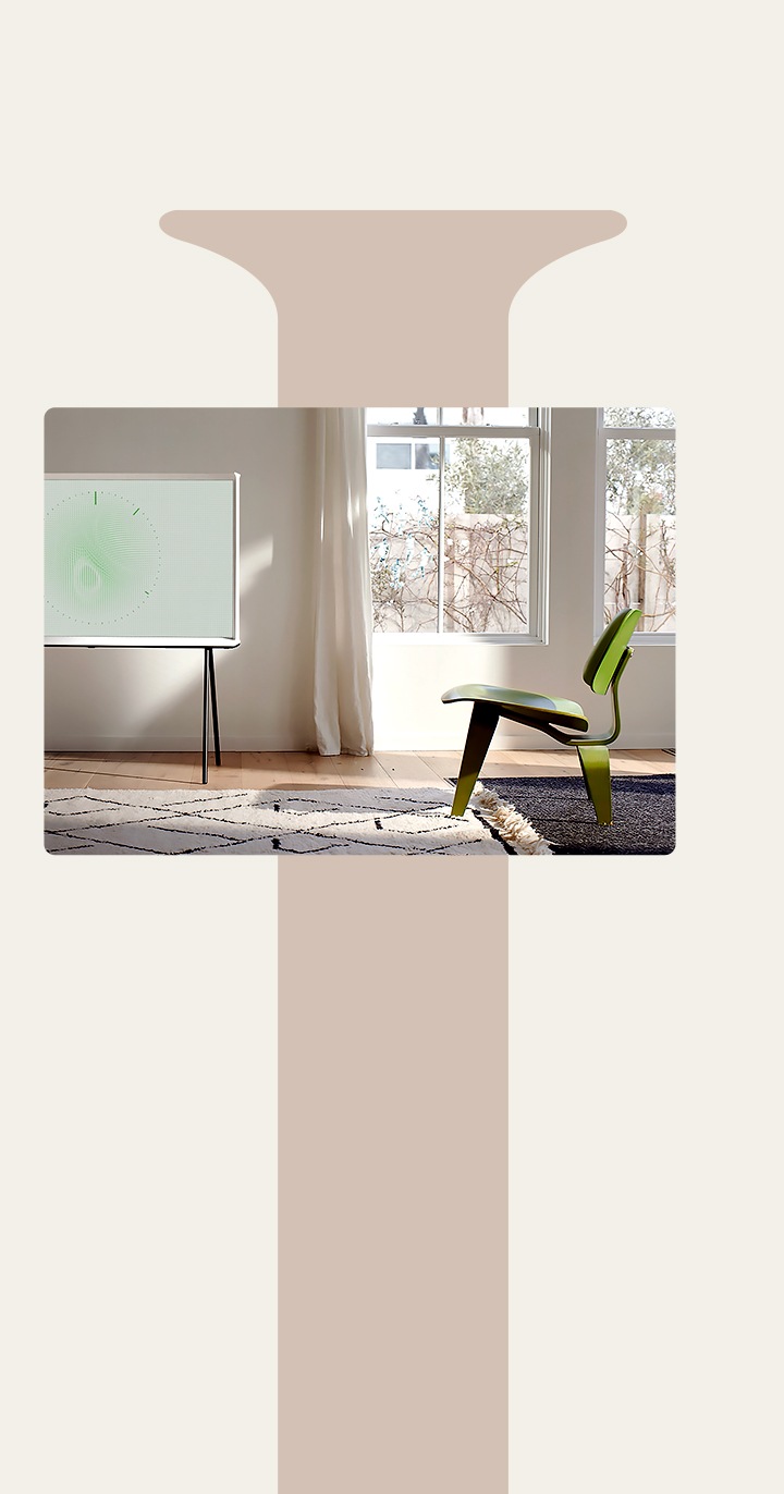 Ein The Serif in Cloud White mit schwarzen Standfuß ist in einem hellen und eleganten Wohnzimmer, mit einem Flächenteppich, einem stilvollen dreibeinigen grünen Stuhl und großen Fenstern. Das Bild auf dem Bildschirm ist im Leaf Ambient Mode in der Farbe Grün.