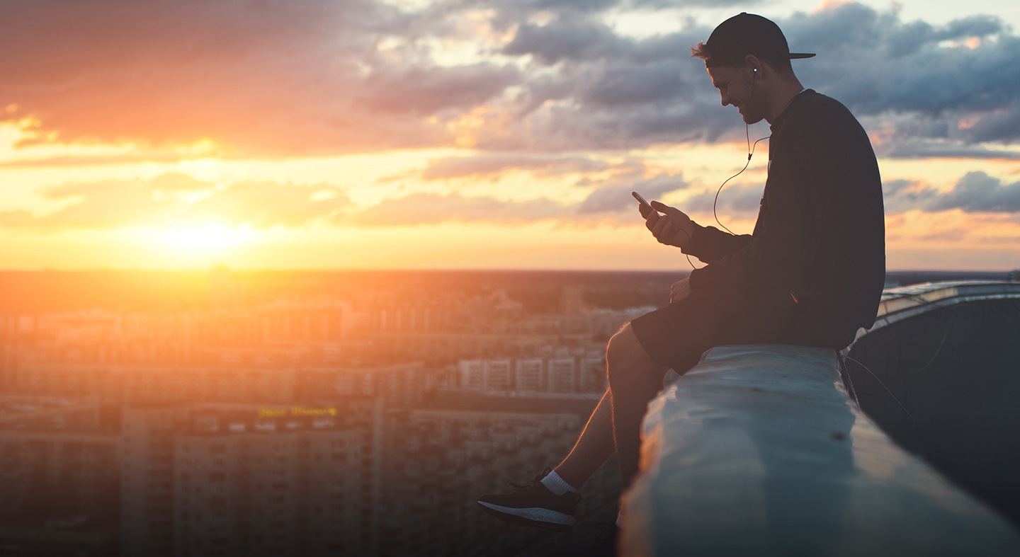 Young man sitting on a high ledge above city smiling and looking at his phone