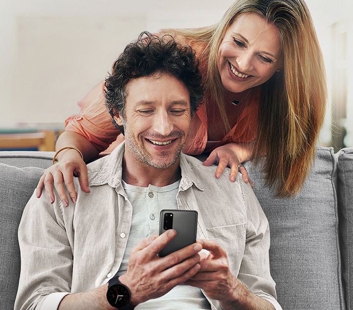 Una pareja de mediana edad más joven que se sonríe. El hombre está sentado en el sofá, sostiene y mira a la pantalla de su Galaxy y la mujer está de pie detrás de él, mirando a la pantalla de Galaxy por encima de su hombro con las manos en los hombros de él.