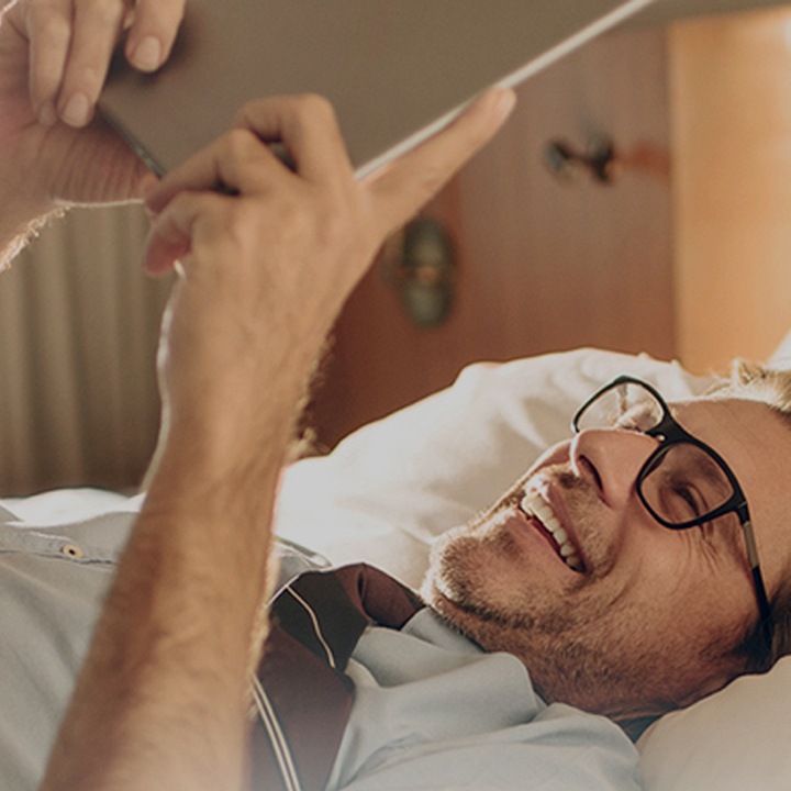 Un homme consulte une tablette Samsung en souriant, allongé sur le lit de sa chambre d'hôtel