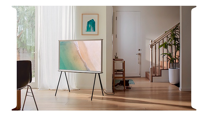 Vidéo design 360° The Serif 2020, une télévision Samsung blanche, est installée sur un pied dans un salon et son écran affiche une plage de sable et une mer transparente, dans une salle de séjour simple et lumineuse avec un plancher en bois et un mur blanc.