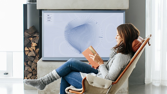Une femme lit un livre, assise dans un fauteuil au milieu du salon. À sa gauche, un modèle bleu coton de la Serif est posé sur un manteau de cheminée. Il est réglé avec le motif Feuille du Mode « Ambient », en couleur bleue.