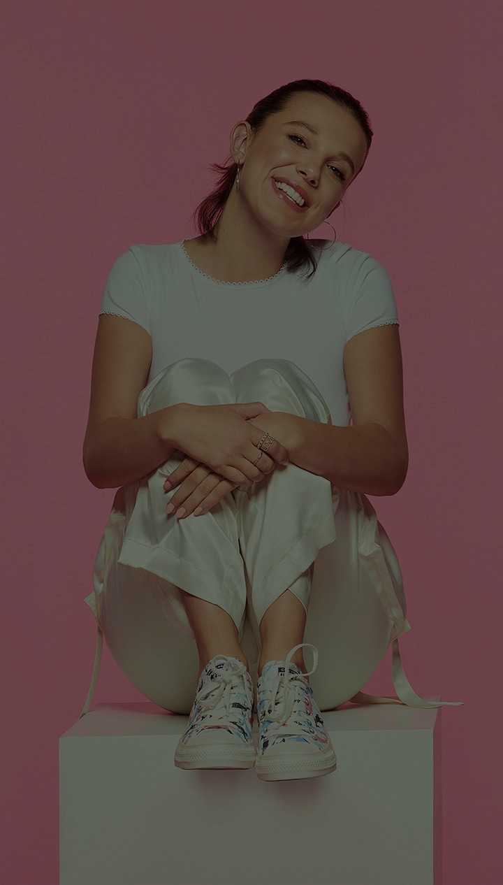Portrait destiné au grand public de Millie Bobby Brown qui pose sur un fond rose. Elle sourit, la tête penchée, assise sur une boîte blanche, les mains croisées sur ses jambes repliées.