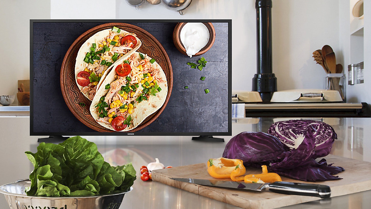 Verduras frescas encima de la encimera de una cocina casera mientras se prepara una receta saludable. Gran pantalla de TV en el fondo, que muestra instrucciones paso a paso de recetas