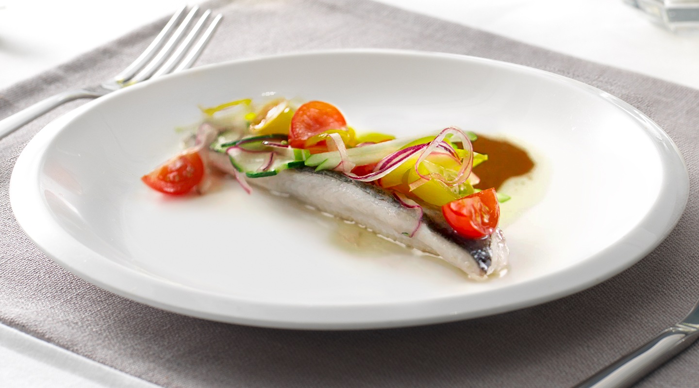 El filete de trucha se presenta con verduras coloridas en la parte superior. El plato blanco está puesto sobre un mantel con cubiertos.