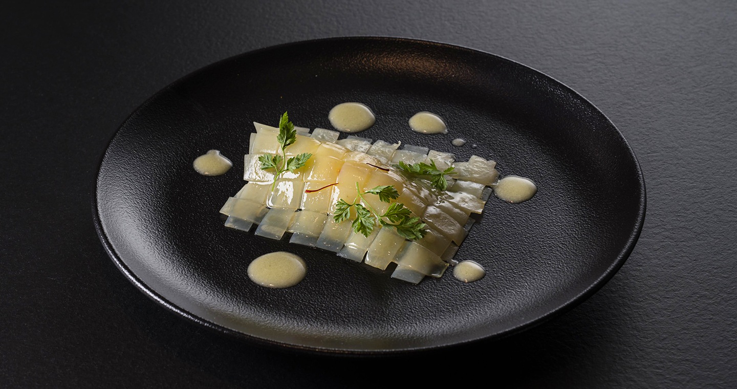 En el centro de un plato negro redondo, se ponen tiras de patata sobre el bacalao que ha sido cuidadosamente colocado. Para completar, se esparce una salsa blanca alrededor del plato, todo acentuado por una pizca de perejil.