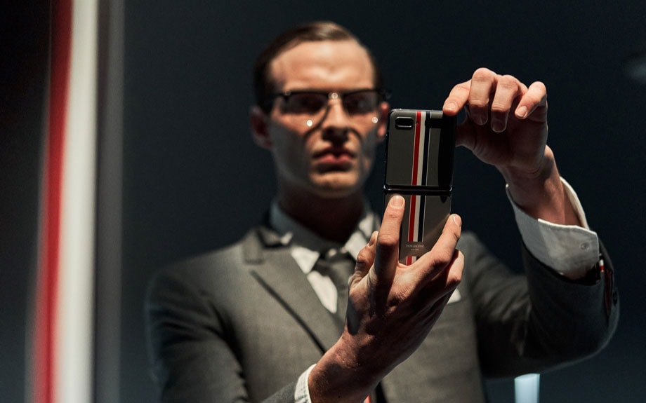 A male model is pictured standing and holding the Galaxy Z Flip Thom Browne Edition unfolded with both hands. He stares intently at the screen.