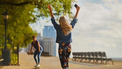 Žena drží obe ruke vysoko nad hlavou s mobilom v pravej ruke. Stojí čelom k osobe blížiacej sa k nej na skateboarde.