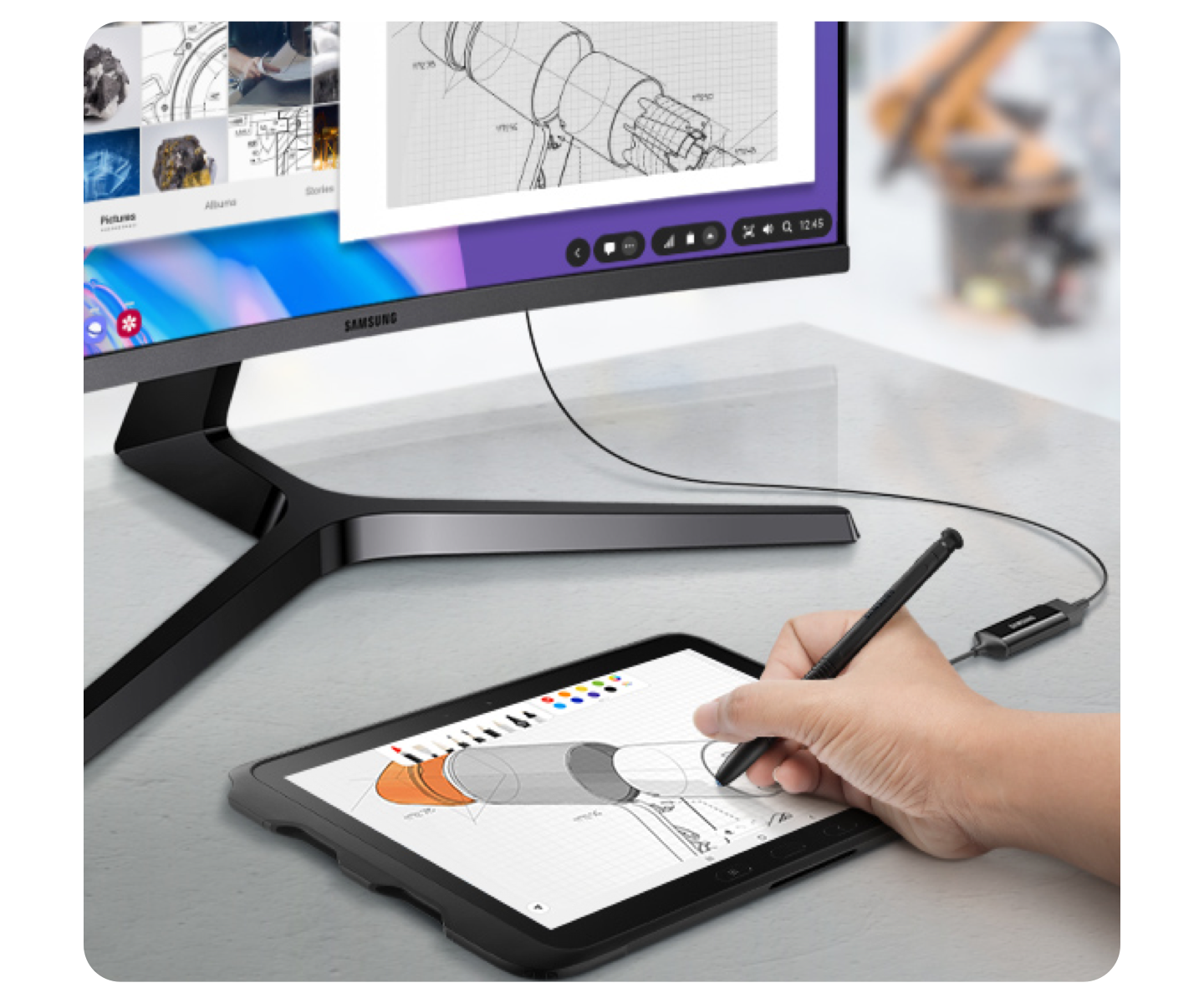 A Samsung curved monitor on a grey desk connected to a Samsung tablet. A hand is using a black S Pen to draw on the tablet.